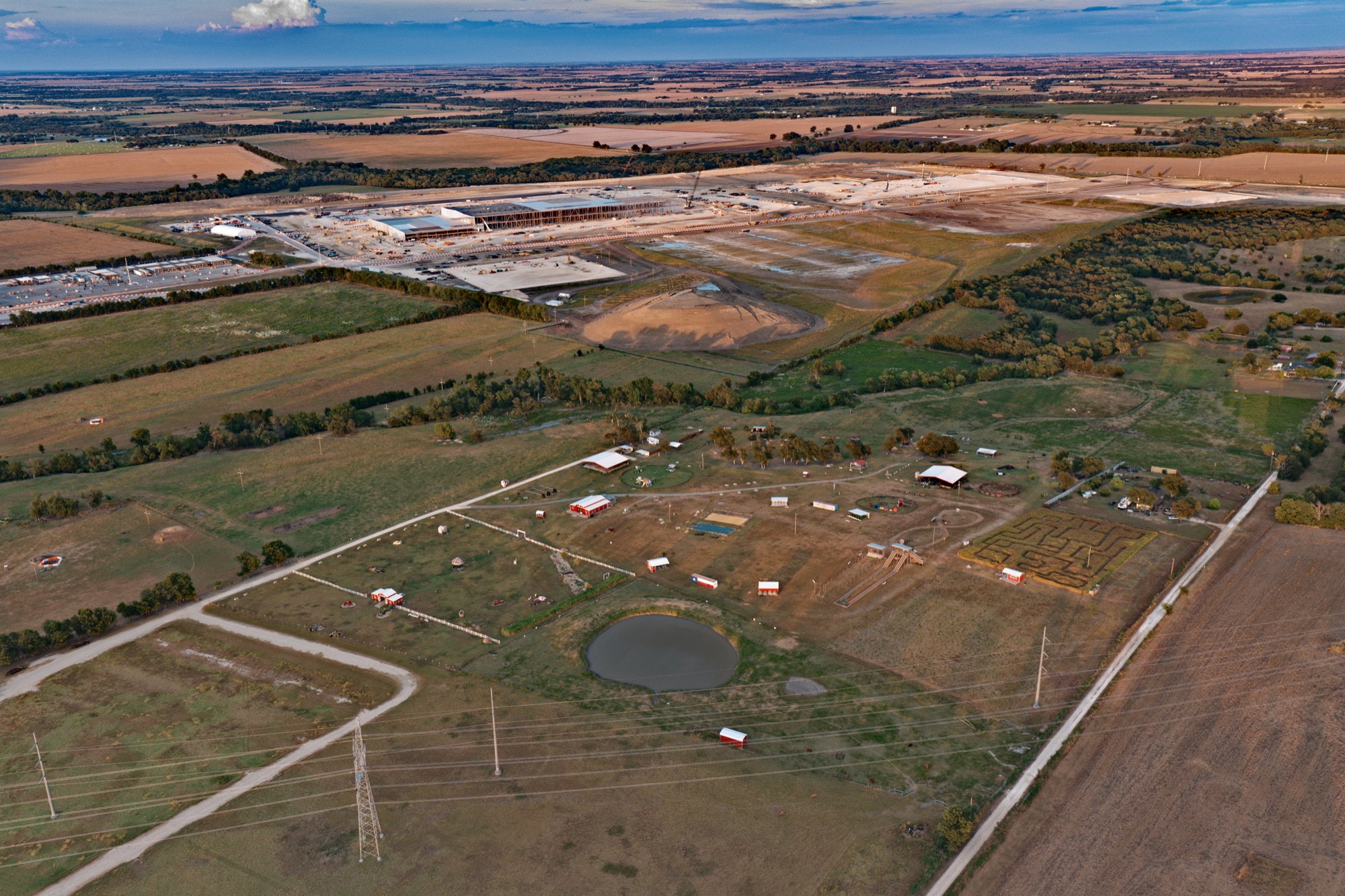 Golden-hour aerial over Bell County: working acreage with a stock pond and barns in the foreground, large-scale construction on the horizon behind it