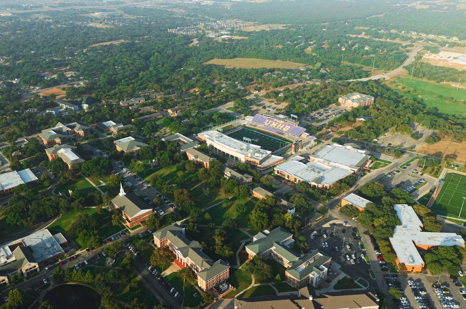 Wide morning aerial of UMHB in Belton, with the stadium, chapel, campus buildings and wooded surroundings.